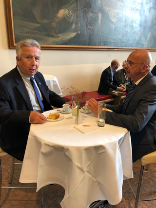 Zwei Männer in Anzügen unterhalten sich an einem Cafétisch mit Kuchen, Kaffee und Wasser, mit einem Gemälde im Hintergrund.