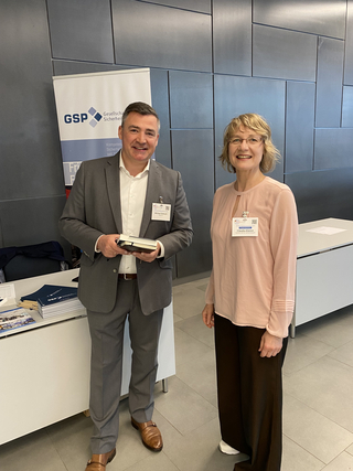 Zwei GSP-Vertreter, Michael Dietsch und Claudia Klemm, lächeln auf einer Konferenz vor einem GSP-Banner und Broschüren.