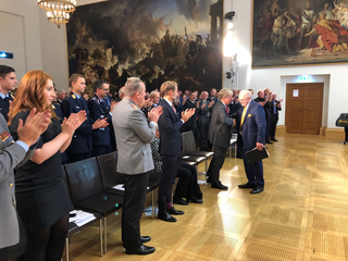 Zwei Männer schütteln Hände in einem großen Saal, umgeben von applaudierendem Publikum, darunter Militärpersonal, vor historischen Gemälden.