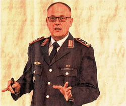 Ein Mann in deutscher Militäruniform mit Brille spricht und gestikuliert mit den Händen bei einer Präsentation.