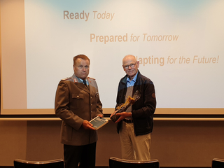Zwei Männer, einer in Uniform mit Buch, der andere mit Geschenk, stehen vor einer Leinwand mit Text zu zukünftiger Bereitschaft.