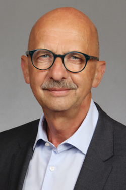 Glatzköpfiger Mann mit Brille und Schnurrbart, in dunklem Anzug und hellblauem Hemd, blickt nach vorne.