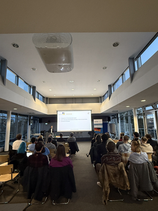 Ein Redner spricht vor einem Publikum in einem Hörsaal. Eine Leinwand zeigt Präsentationsfolien über John F. Kennedy und die Kubakrise.