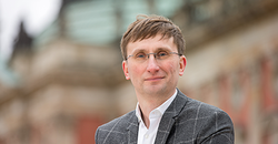 Lächelnder Mann mit Brille in karierter Jacke, blickt in die Kamera, verschwommenes Gebäude im Hintergrund.