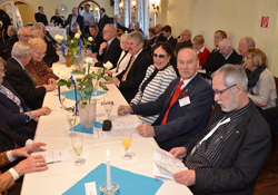 Viele Gäste bei einer festlichen Veranstaltung, sitzend an langen Tischen, dekoriert mit weißen Rosen und Kerzen. Manche unterhalten sich.