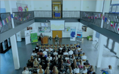 Schüler sitzen in einer Aula und lauschen einem Vortrag eines Mannes. Im Hintergrund Projektion, Podium, und EU-Flaggenkette.
