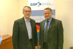 Zwei Männer in Anzügen lächeln vor einem GSP-Banner (Gesellschaft für Sicherheitspolitik) mit Text zu Frieden und Sicherheit.