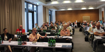 Eine Frau spricht in ein Mikrofon an einem Konferenztisch, während ein Publikum aus meist älteren Erwachsenen zuhört.