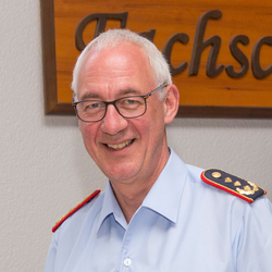 Ein lächelnder Mann mit grauen Haaren und Brille in einem hellblauen Uniformhemd mit verzierten Schulterklappen.
