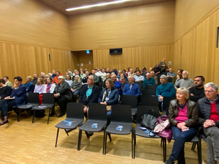 Eine große Gruppe Erwachsener sitzt in einem Auditorium mit Holzwänden und verfolgt eine Präsentation oder Veranstaltung.