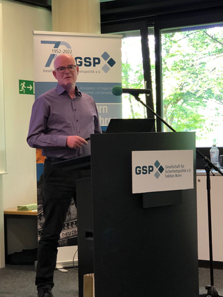Ein Mann im lila Hemd mit Brille präsentiert auf einer GSP-Veranstaltung hinter einem Rednerpult mit dem GSP Bonn Logo.