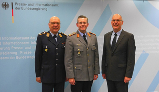 Drei lächelnde Männer, zwei in Uniform und einer im Anzug, vor dem Logo des Presse- und Informationsamtes.