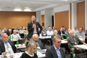Ein Redner spricht vor einem Raum voller Zuhörer, die bei einer Konferenz oder Tagung an Tischen sitzen.