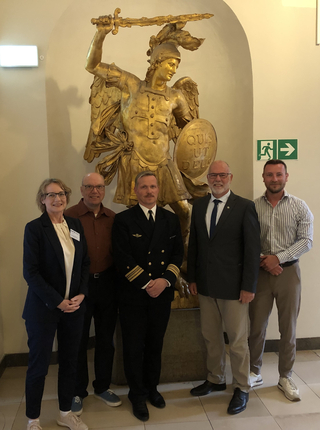 Fünf Personen, darunter eine in Uniform, posieren vor einer großen goldenen Statue des Erzengels Michael mit Schwert und Schild.