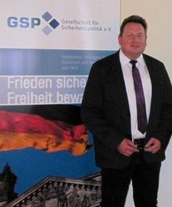 Mann im Anzug vor GSP-Banner mit Deutschlandflagge und Text 'Frieden sichern, Freiheit bewahren'.
