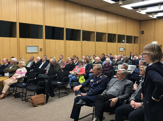 Zuschauer sitzen in einem holzvertäfelten Konferenzraum bei einer Präsentation; eine Frau steht im Vordergrund.