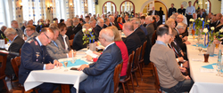 Große Gruppe von Menschen, meist ältere Männer, sitzen an Tischen in einer Halle mit Dekorationen wie weißen Rosen und blauen Bändern.