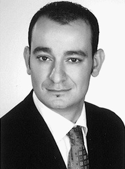 Monochromes Porträt eines Mannes im Anzug mit gemusterter Krawatte, kurzen dunklen Haaren und Lippenbart, lächelnd.