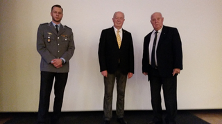 Drei Männer stehen nebeneinander vor einem hellen Hintergrund: einer in Militäruniform und zwei in dunklen Anzügen.
