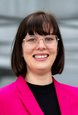 Lächelnde Frau mit kurzen braunen Haaren, goldener Brille und leuchtend pinkfarbenem Blazer, auf einer grauen Leiste lehnend.