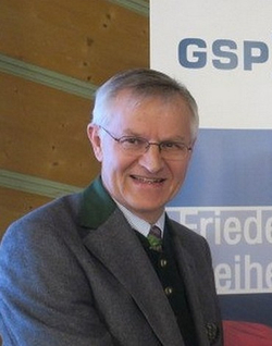 Lächelnder älterer Mann mit Brille und Trachtenjanker vor einem Hintergrund mit "GSP" und "Frieden Freiheit".