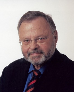 Kopfaufnahme eines älteren Mannes mit Brille, grauem Bart, blauem Hemd und gestreifter Krawatte, blickt in die Kamera.