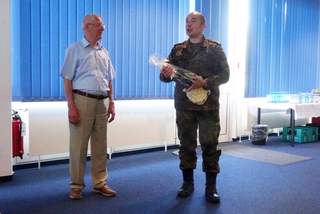 Ein Mann in Militäruniform hält ein Geschenk und spricht mit einem älteren Mann in hellblauem Hemd in einem Raum.