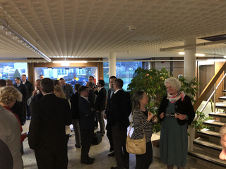 Menschen bei einem gesellschaftlichen Anlass in einem hellen Foyer mit großen Fenstern und Treppe.