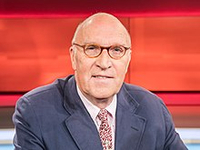 Nahaufnahme des österreichischen Schauspielers Peter Weck, ein Glatzkopf mit Brille, blauem Sakko und gemusterter Krawatte, lächelt vor rotem Hintergrund.