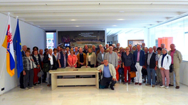 Eine große Gruppe Erwachsener posiert in einem modernen Saal mit Maastricht- und EU-Flaggen und einer Tafel zum Vertrag von Maastricht.