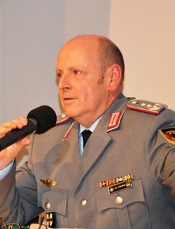 Mann in deutscher Militäruniform mit Medaillen und Rangabzeichen spricht ins Mikrofon.