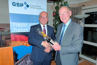Zwei Männer in Anzügen schütteln Hände, einer überreicht eine Geschenkflasche vor einem GSP-Banner.