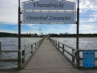 Langer Holzsteg mit Schild „Vinetabrücke Ostseebad Zinnowitz“, führt zu einem Strand mit Gebäuden.