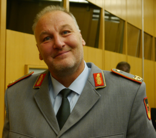 Ein lächelnder Mann in grauer deutscher Militäruniform mit roten und goldenen Abzeichen blickt in die Kamera.