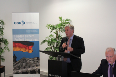 Mann spricht an einem Pult bei einer sicherheitspolitischen Veranstaltung, mit GSP-Banner und Reichstagsbild im Hintergrund.
