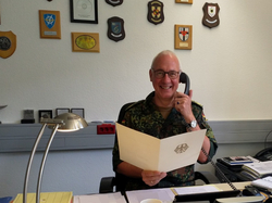 Ein lächelnder Mann in Tarnuniform sitzt am Schreibtisch, telefoniert und hält ein Dokument mit dem Bundesadler.