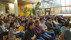 Große Gruppe junger, leger gekleideter Menschen, wohl Studenten, sitzt in Reihen in hellem Raum mit gelben Wänden und Fenstern.