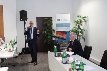 Mann spricht mit Mikrofon bei einer Konferenz; ein Zuhörer sitzt am Tisch. Im Hintergrund Banner der Gesellschaft für Sicherheitspolitik.