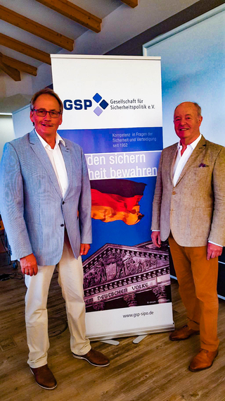 Zwei lächelnde Männer in Sakkos vor einem GSP-Banner der Gesellschaft für Sicherheitspolitik mit Reichstag und Flagge.