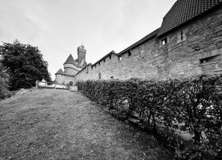Schwarz-Weiß-Ansicht einer alten Burgmauer, Hecke, grasbewachsenem Hang und fernen Türmen unter hellem Himmel.