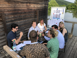 Eine Gruppe diskutiert an einem Holztisch im Freien am See, mit Flipchart und Schwan im Hintergrund.