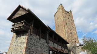 Historische Steinburgmauer mit Holzgang und einem hohen, schmalen Turm mit Giebel, unter bewölktem Himmel.