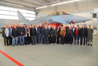 Eine große Gruppe meist älterer Erwachsener posiert vor einem deutschen Eurofighter Typhoon Jet in einem Hangar.