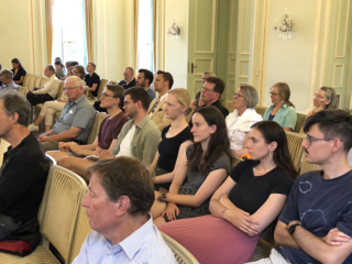 Vielfältiges Publikum aus Männern und Frauen unterschiedlichen Alters sitzt in Stuhlreihen und blickt nach vorne in einem Saal.