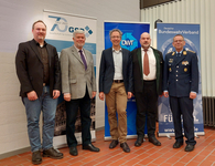 Fünf Männer, darunter einer in Militäruniform, stehen vor drei Bannern deutscher Organisationen.