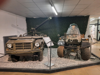Zwei historische Militärfahrzeuge: ein Geländewagen mit rückstoßfreiem Geschütz und ein Spezialfahrzeug mit Mannequin, im Museum.