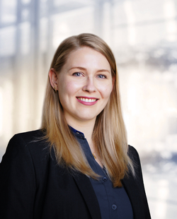 Professionelles Porträt einer lächelnden blonden Frau mit blauen Augen in einem schwarzen Blazer und blauen Hemd.