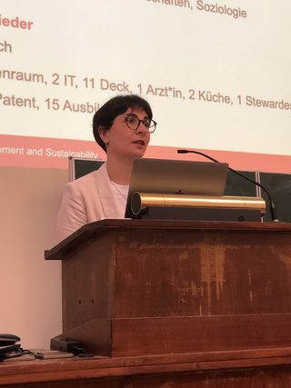 Eine Frau mit Brille und dunklem Haar spricht an einem Holzpult, dahinter eine Präsentationsfolie.