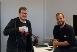 Zwei lächelnde Männer in einer Büroumgebung. Ein Mann mit Brille hält eine Tasse, der andere mit Bart sitzt auf einem Tisch.