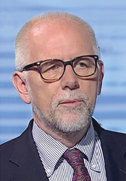 Kopfaufnahme eines kahlköpfigen Mannes mit grauem Bart und schwarzer Brille, gestreiftem Hemd und Krawatte vor blauem Hintergrund.